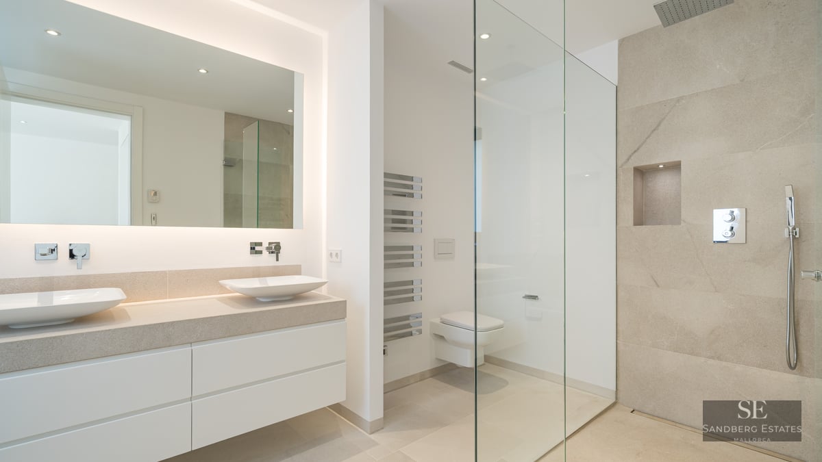 Modern master bathroom featuring a double white vanity, large backlit mirror, and a walk-in glass shower with stone tiles.