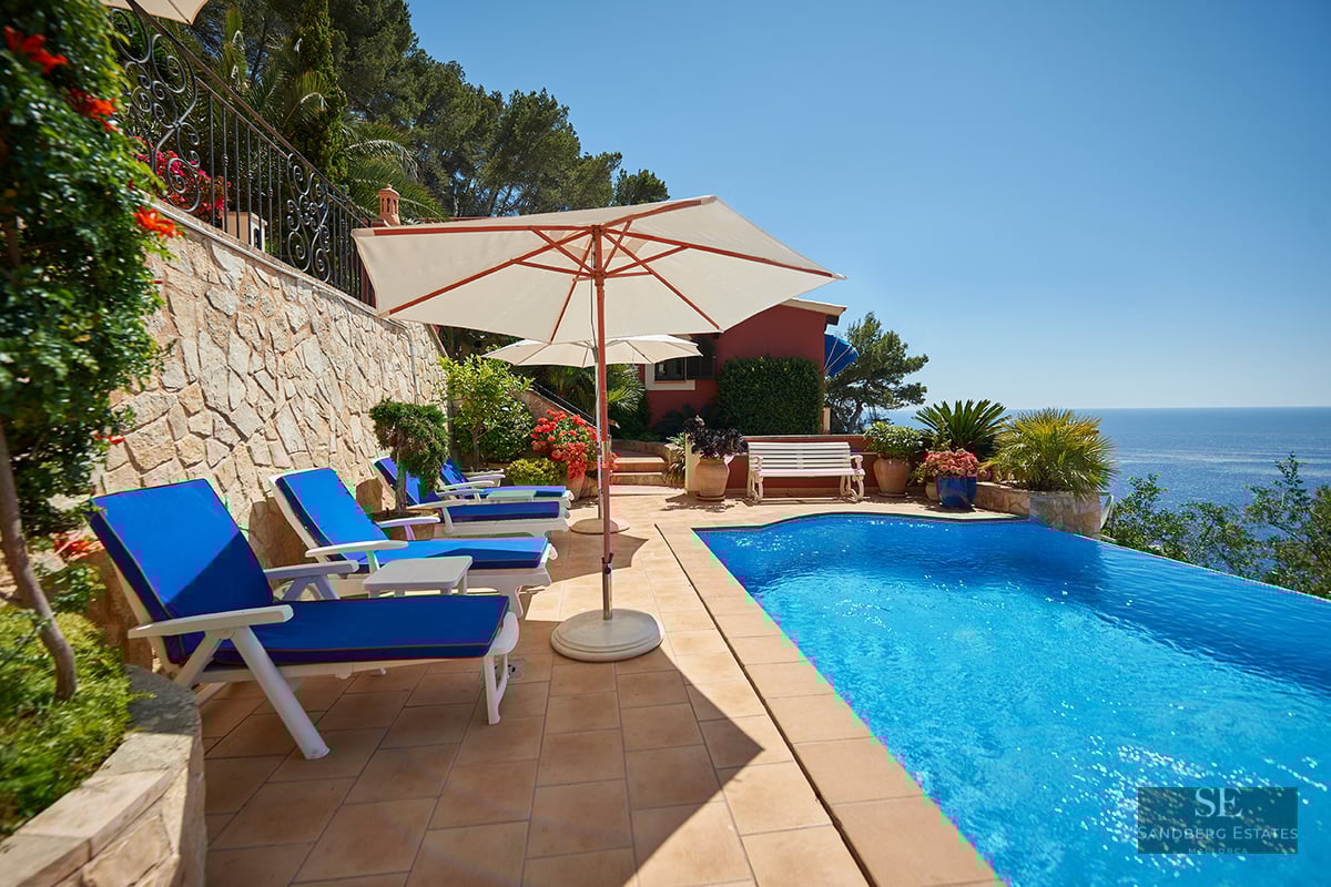 Azure infinity pool on a terracotta terrace with blue loungers and white umbrellas overlooking the Mediterranean Sea.