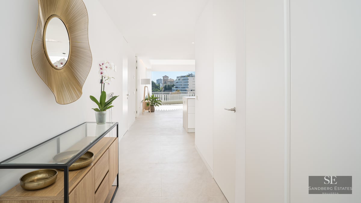 White hallway with gold sunburst mirror and wooden console, leading towards a sunny city view.