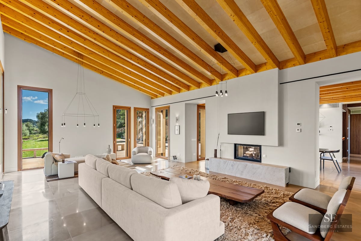 Spacious modern living room featuring high ceilings with wood beams, a large sofa, fireplace, and garden views.