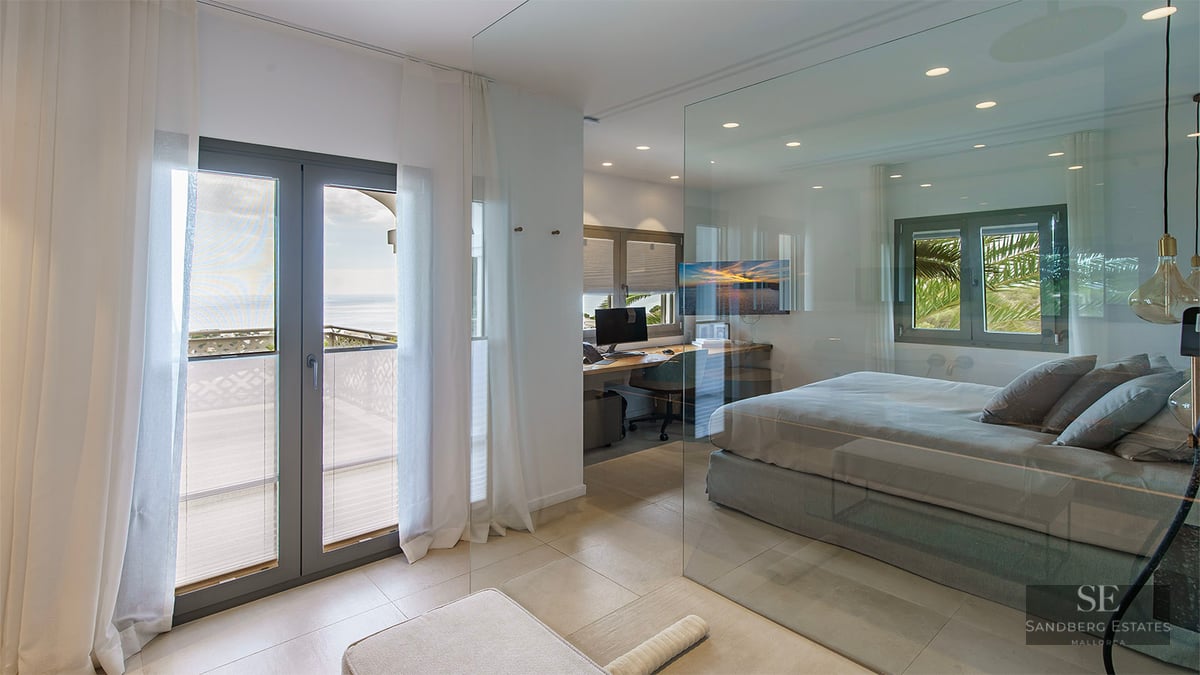 Modern bedroom with floor-to-ceiling glass wall, integrated office area, and balcony overlooking the sea.