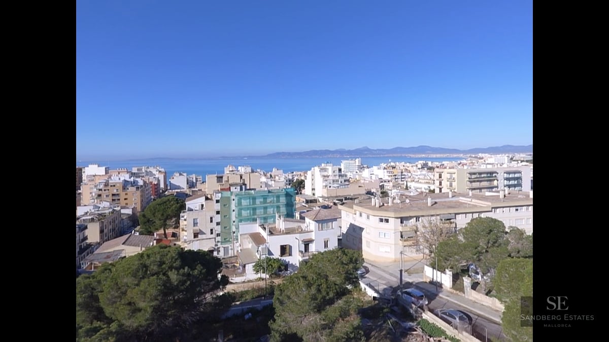 Elevated view of a Mediterranean coastal city with white buildings, green trees, and the blue sea on the horizon.