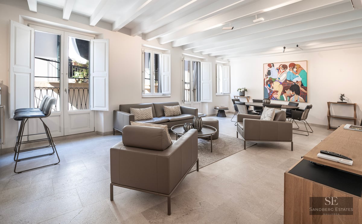 Salon lumineux avec canapés en cuir gris, plafond aux poutres blanches et coin repas avec une œuvre d'art colorée.