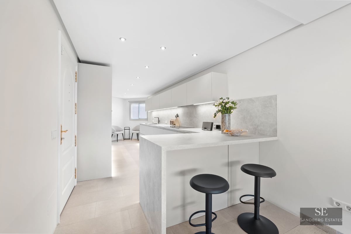 Une cuisine blanche moderne avec un comptoir en pierre, deux tabourets noirs et un éclairage LED intégré.