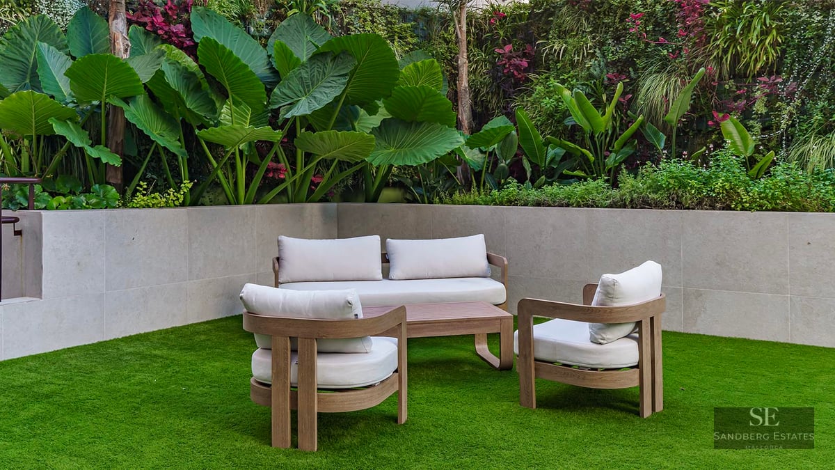 Modern wood patio furniture on green grass in front of a stone wall and lush tropical plants.