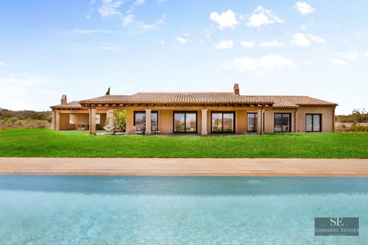 Large turquoise pool in front of a modern Mediterranean villa with a green lawn and terracotta roof under a blue sky.