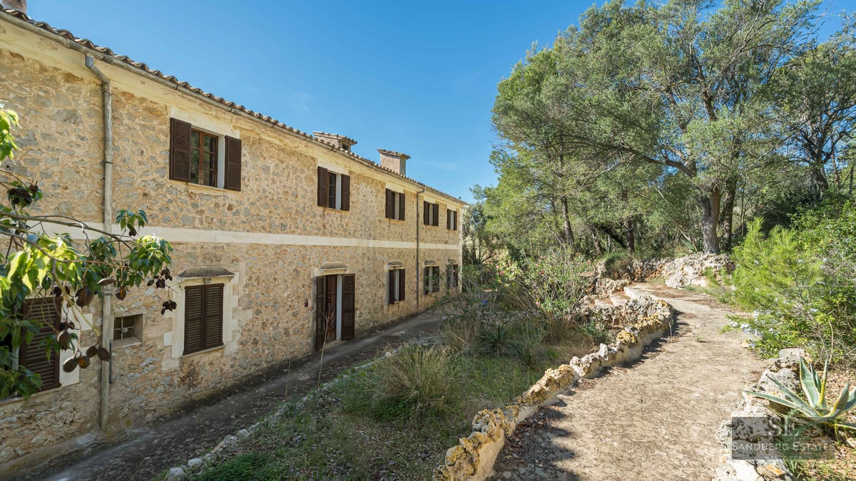 A two-story rustic stone building with dark brown shutters surrounded by lush green trees under a clear blue sky.