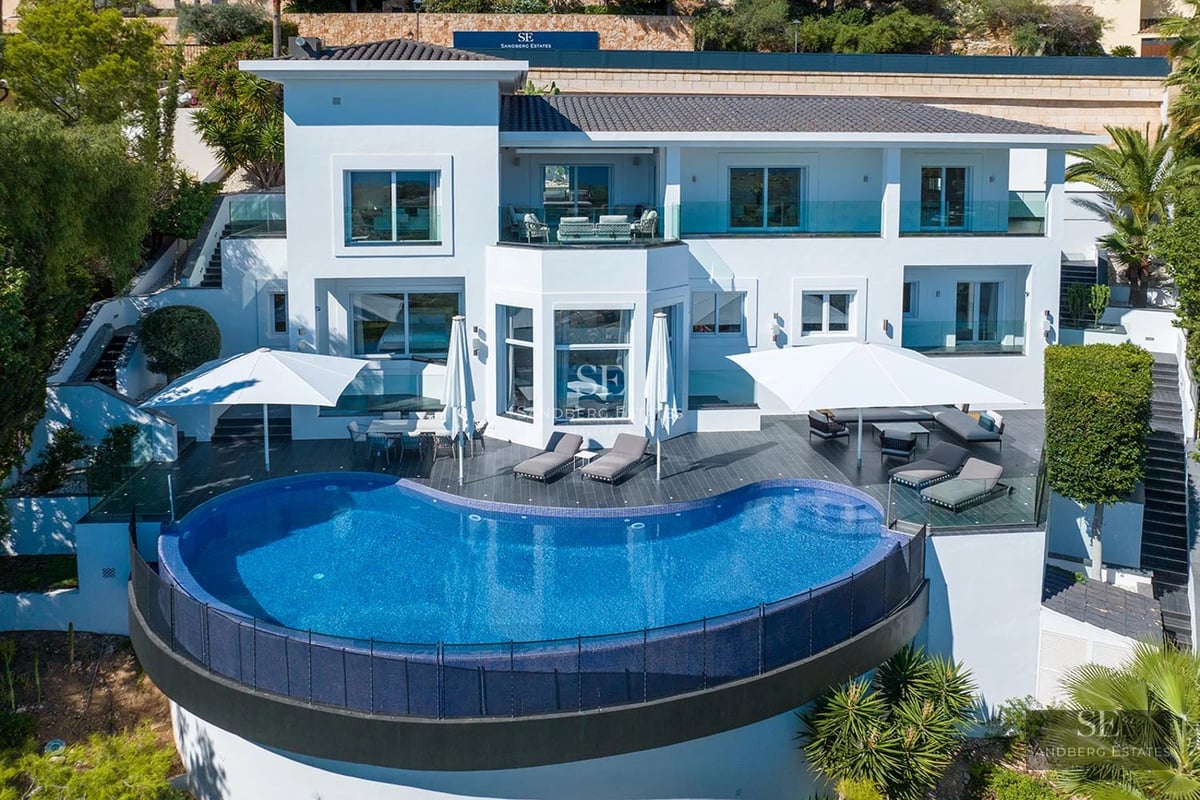 A multi-level white modern villa with a large curved infinity pool, expansive dark gray terrace, and white sun umbrellas.