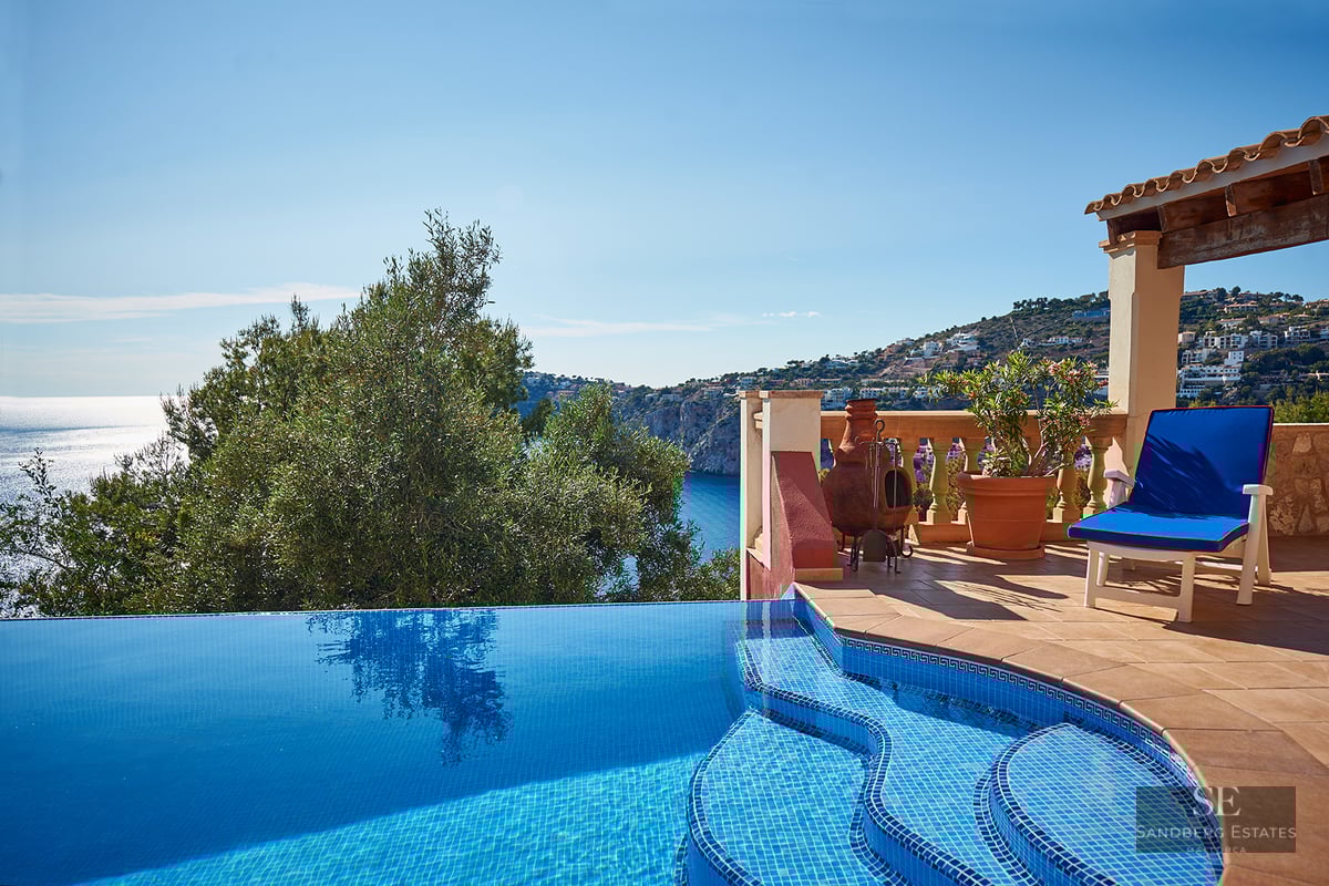 Infinity pool with blue mosaics next to a terracotta terrace overlooking the Mediterranean sea and coastline.
