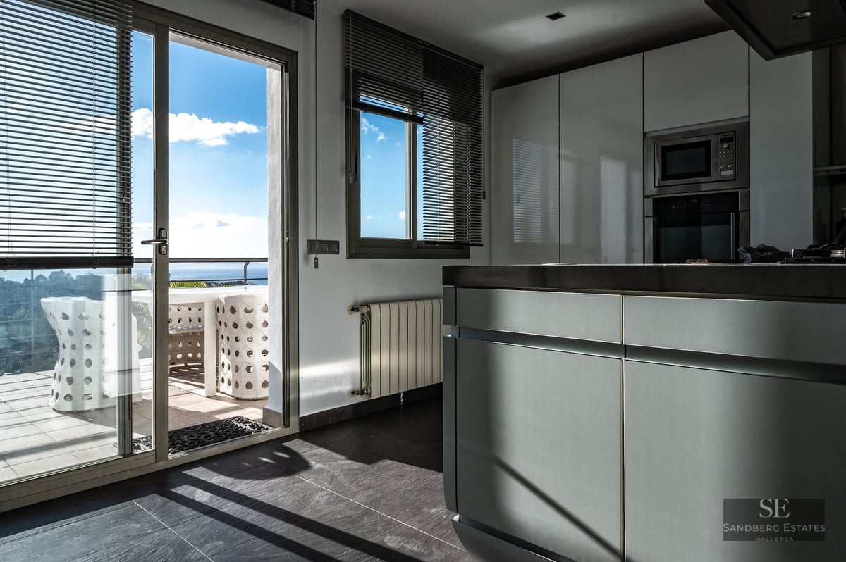 Minimalist white kitchen with an island and large sliding glass doors opening to a sunny terrace with sea views.