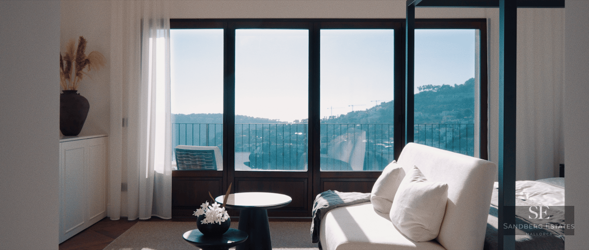 Modern master bedroom featuring a white sofa, canopy bed, and floor-to-ceiling windows overlooking the sea and hills.