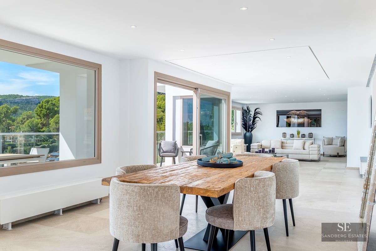 A bright, open-plan interior featuring a rustic wood dining table, beige chairs, and large windows with mountain views.