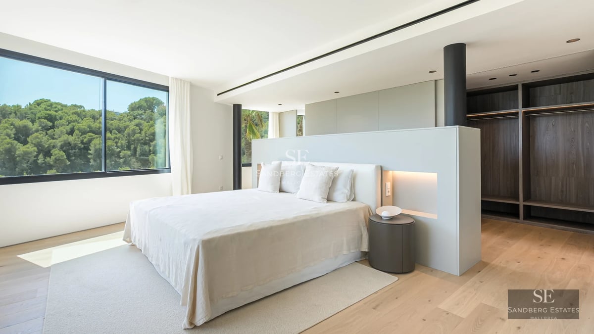 Spacious living room with floor-to-ceiling windows, light wood floors, and a fireplace. Minimalist furniture and abundant natural light. Garden view.