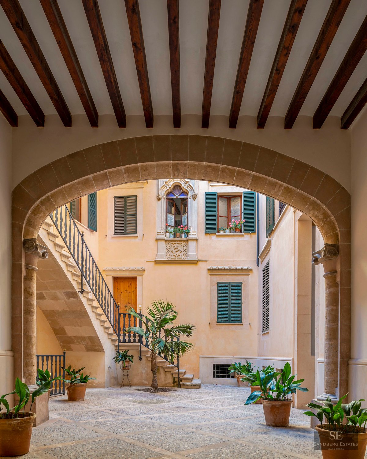Interior courtyard featuring a stone arch, wooden beams, stone staircase, and potted plants under natural light.