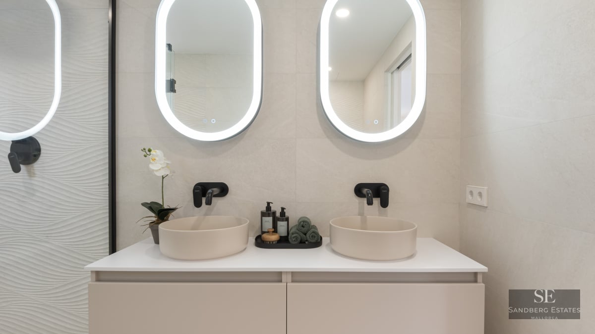 Modern bathroom with two round vessel sinks, black faucets, and oval backlit mirrors on a beige wall.