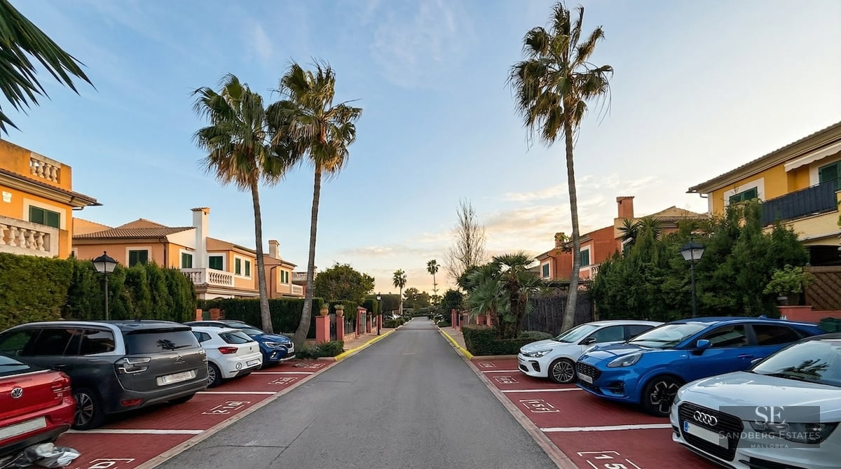 A residential street lined with palm trees, Mediterranean-style houses, and marked parking stalls with cars.
