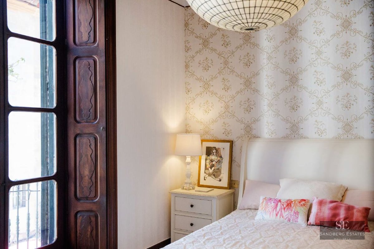 Bedroom featuring patterned wallpaper, a white bed, bedside lamp, and a dark wooden window frame.