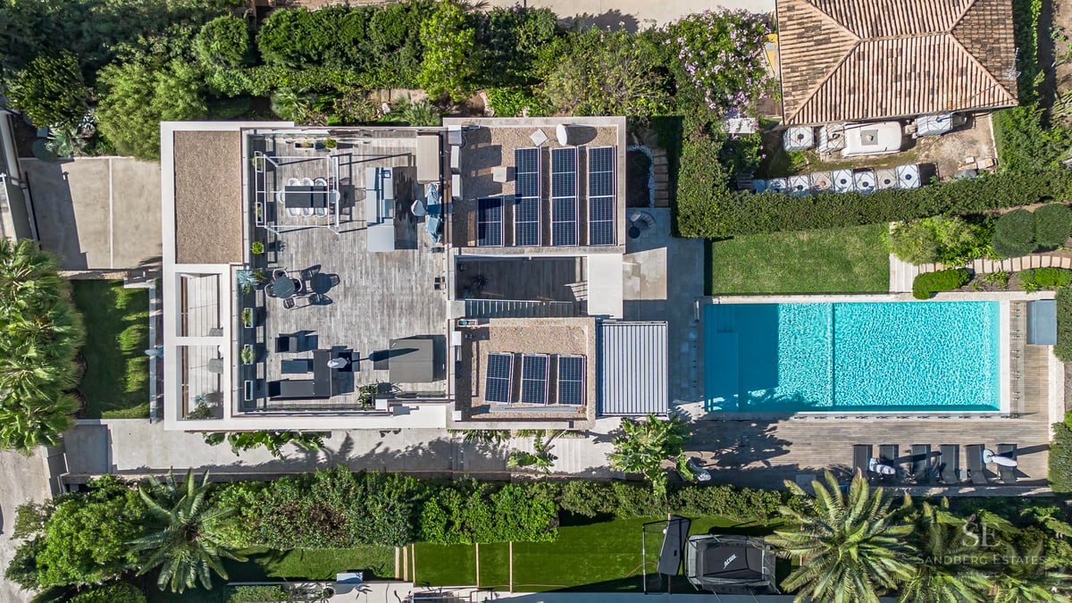 Vue plongeante d'une villa moderne avec terrasse en bois, panneaux solaires et piscine entourée de verdure.