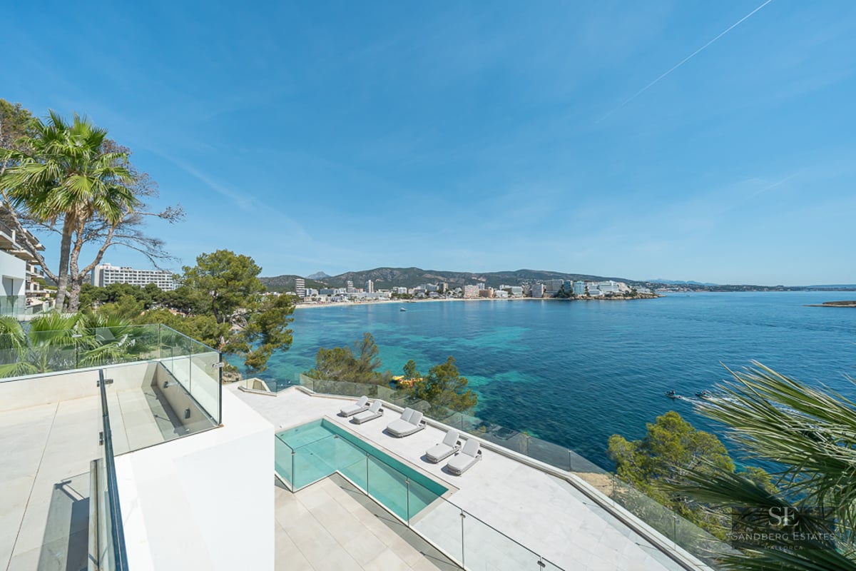 Modern white terrace with infinity pool and glass railings overlooking a turquoise bay and coastline.