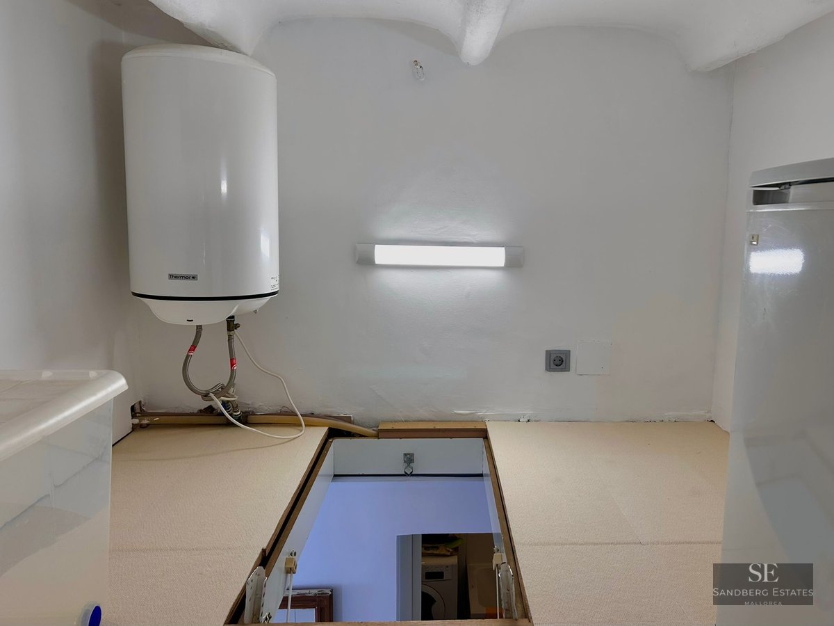 White utility room with an electric water heater on the wall and a rectangular hatch open to the floor below.