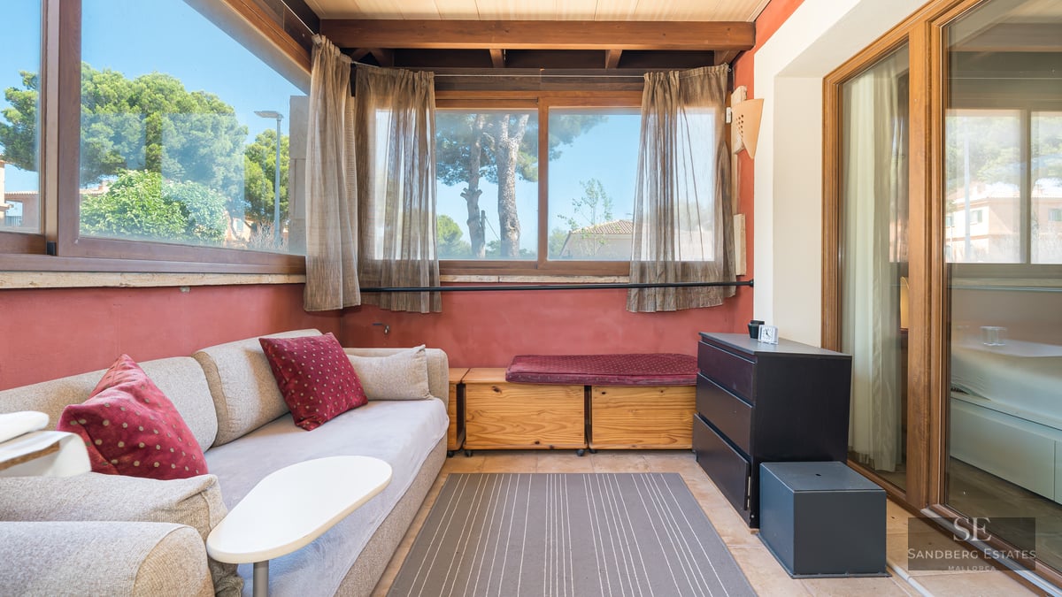 Sunlit living area with beige sofa, wooden beams, terracotta walls, and large windows overlooking green trees.