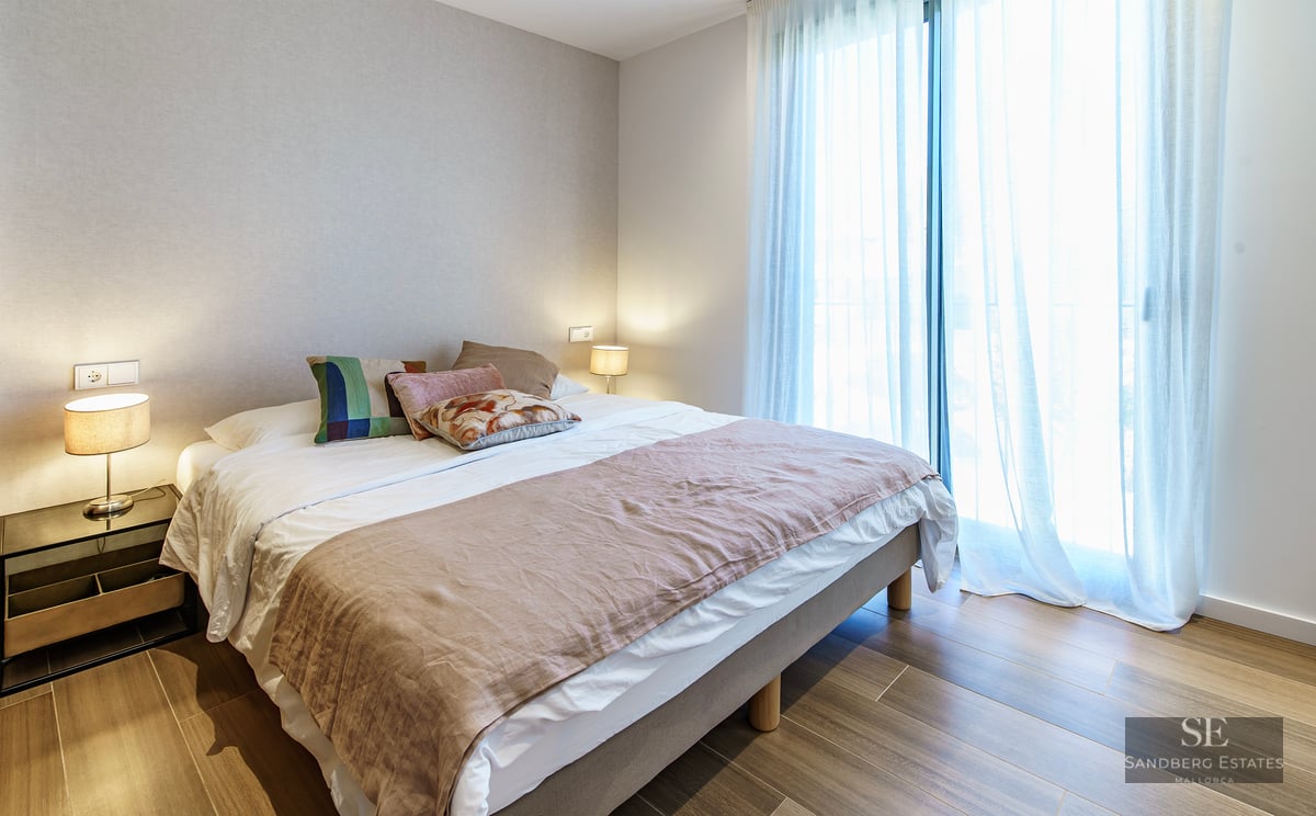 Bright bedroom with a large bed, decorative pillows, bedside lamps, and floor-to-ceiling windows with sheer white curtains.