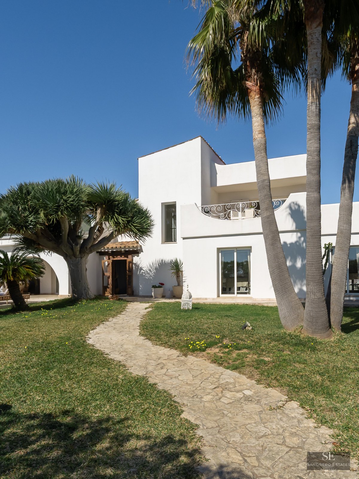 White Mediterranean villa with tall palm trees, a dragon tree, and a stone path on a sunny day.
