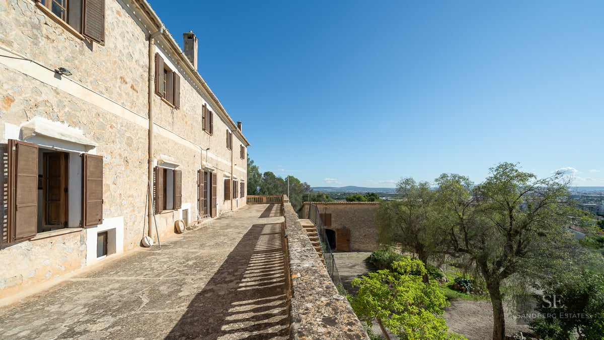 A long stone terrace on a rustic building with wooden shutters, overlooking a green landscape under a clear blue sky.