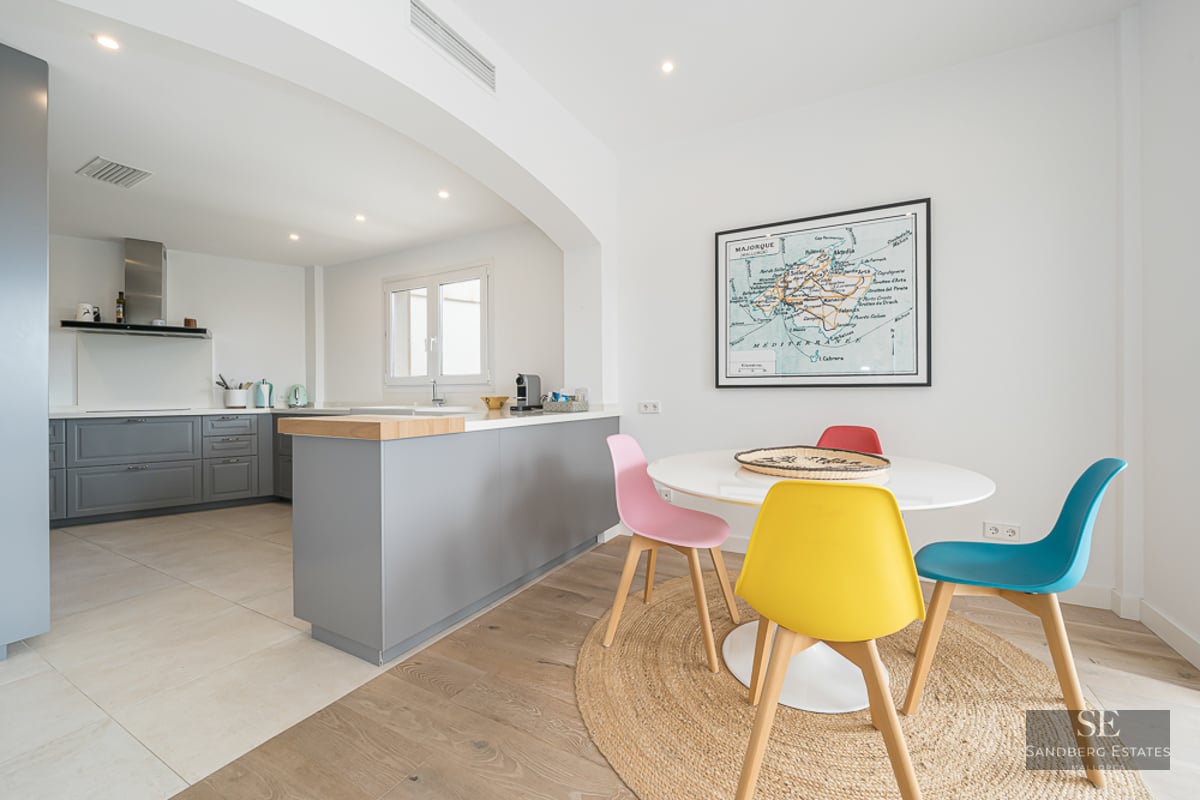 Salle à manger lumineuse avec table ronde blanche, chaises colorées et tapis en jute à côté d'une cuisine grise.