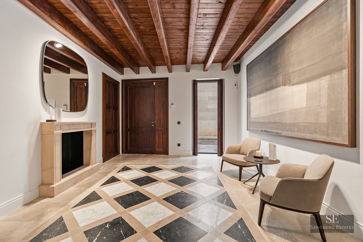 Luxury entry with marble checkerboard floor, exposed wood ceiling beams, stone fireplace, and designer armchairs.