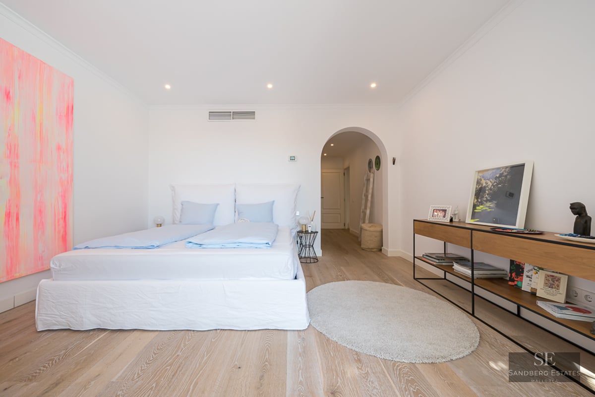 Chambre lumineuse avec murs blancs, parquet clair, grand lit avec draps bleus et passage voûté vers le couloir.