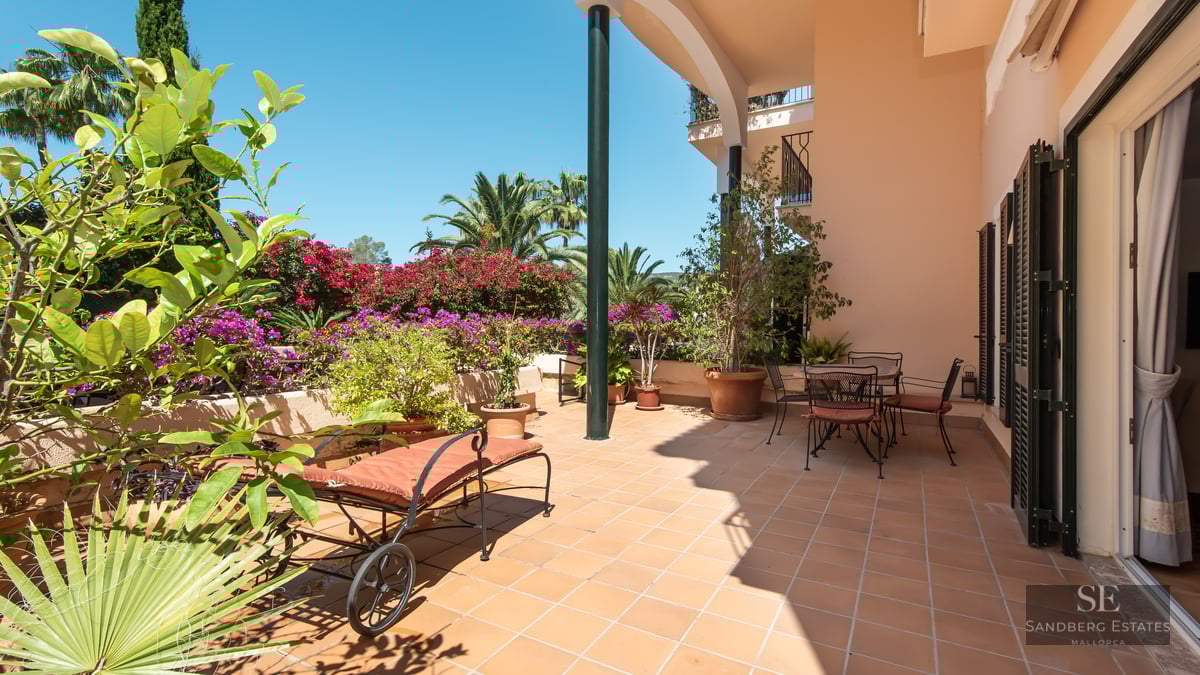 Terracotta terrace with a sun lounger and dining area surrounded by bougainvillea and palm trees under a blue sky.