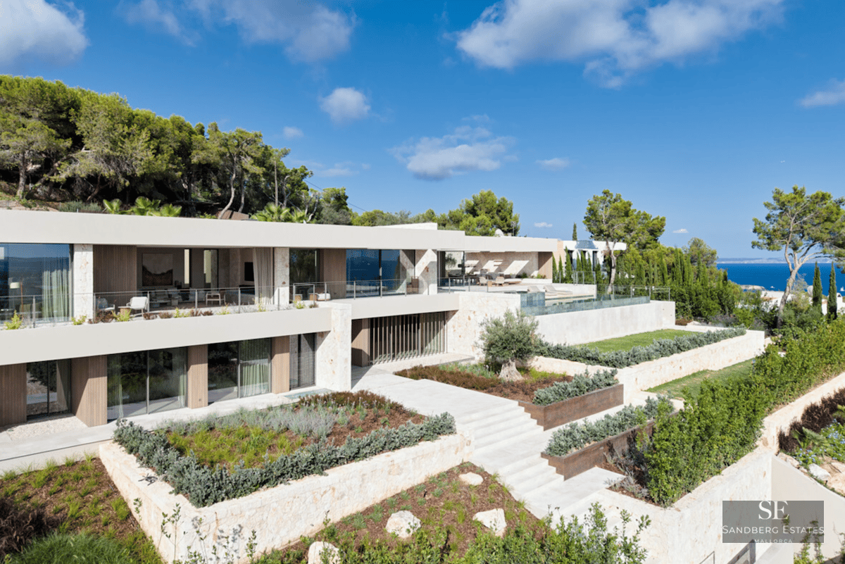 Modern two-story luxury villa with stone facades, glass balconies, and tiered Mediterranean gardens overlooking the sea.