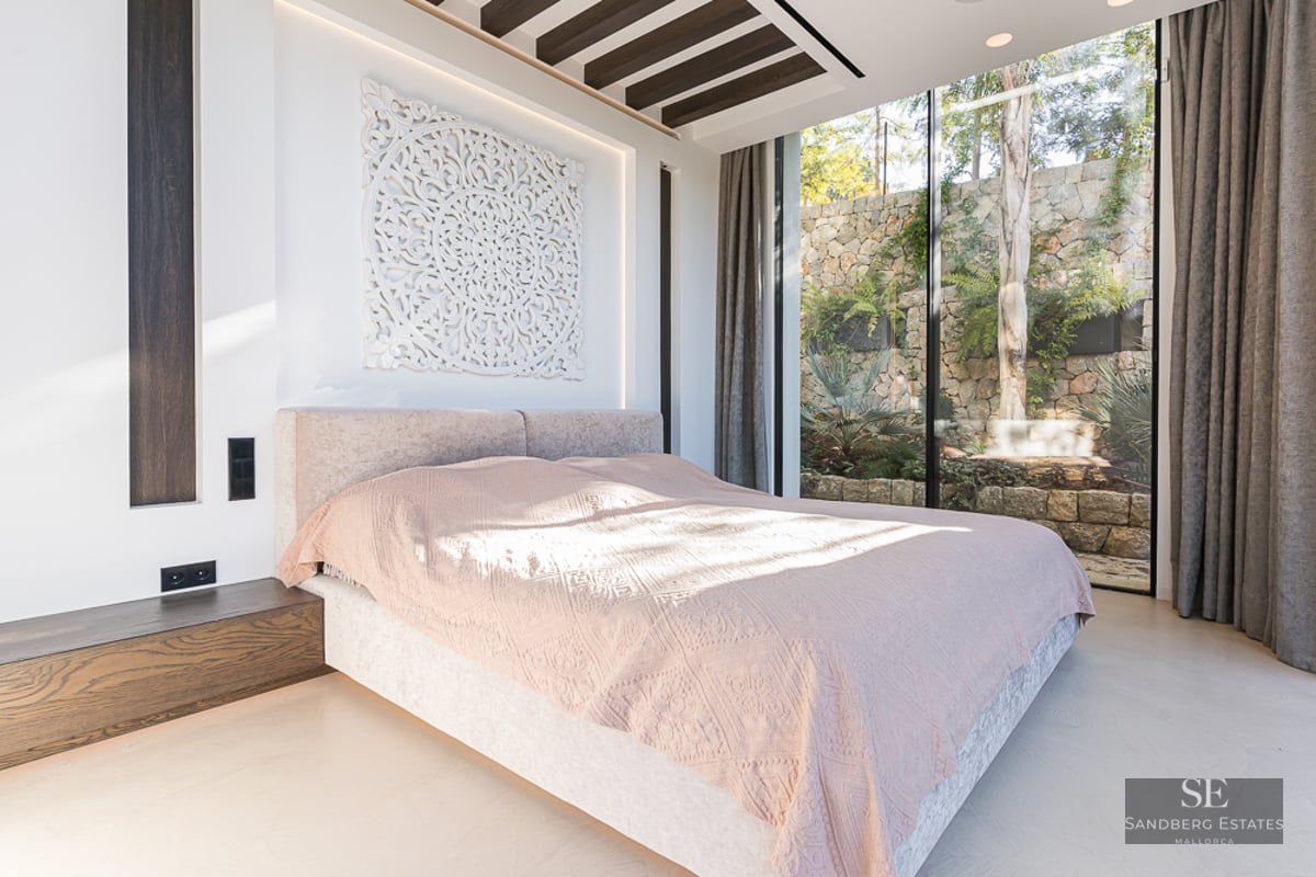 Bright master bedroom with a large bed, intricate wall art, and floor-to-ceiling windows looking out to a stone garden wall.