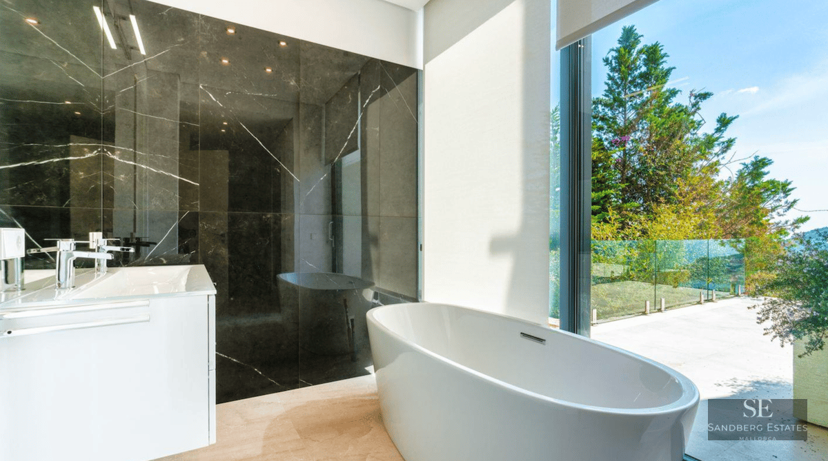 Modern master bathroom featuring a white freestanding bathtub next to a floor-to-ceiling window with green garden views.
