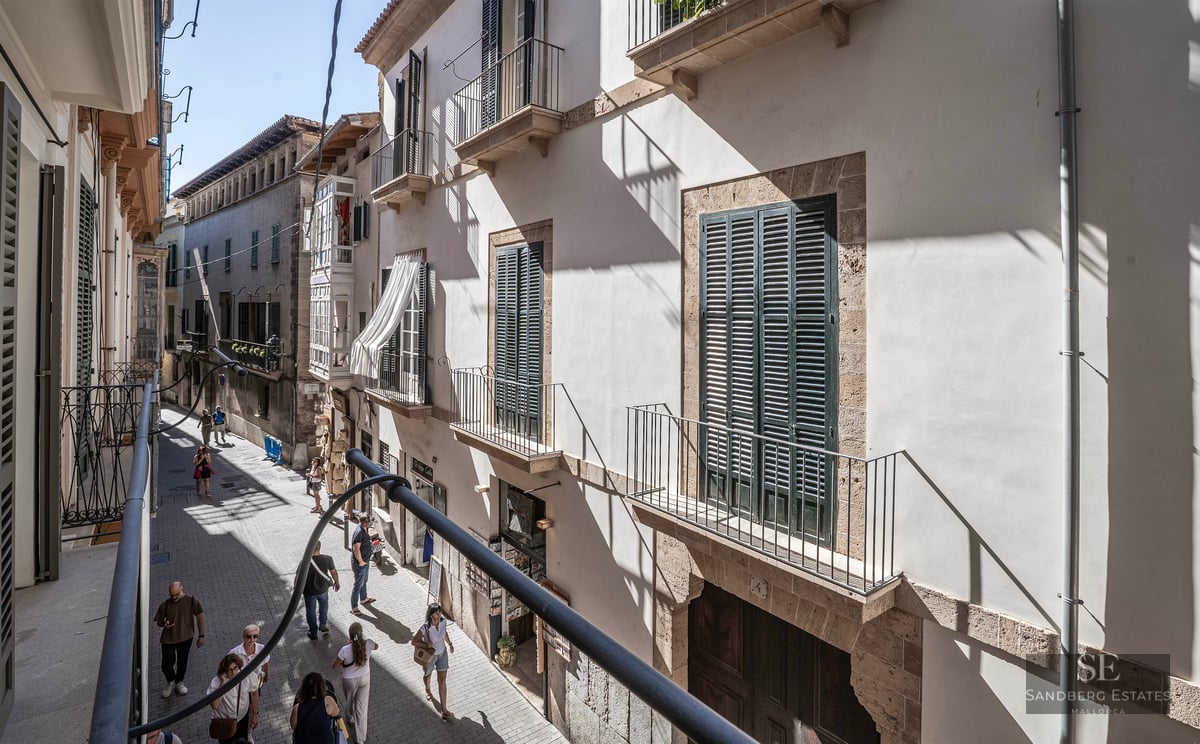 View from a balcony looking down a narrow street with light stone buildings, green shutters, and people walking.