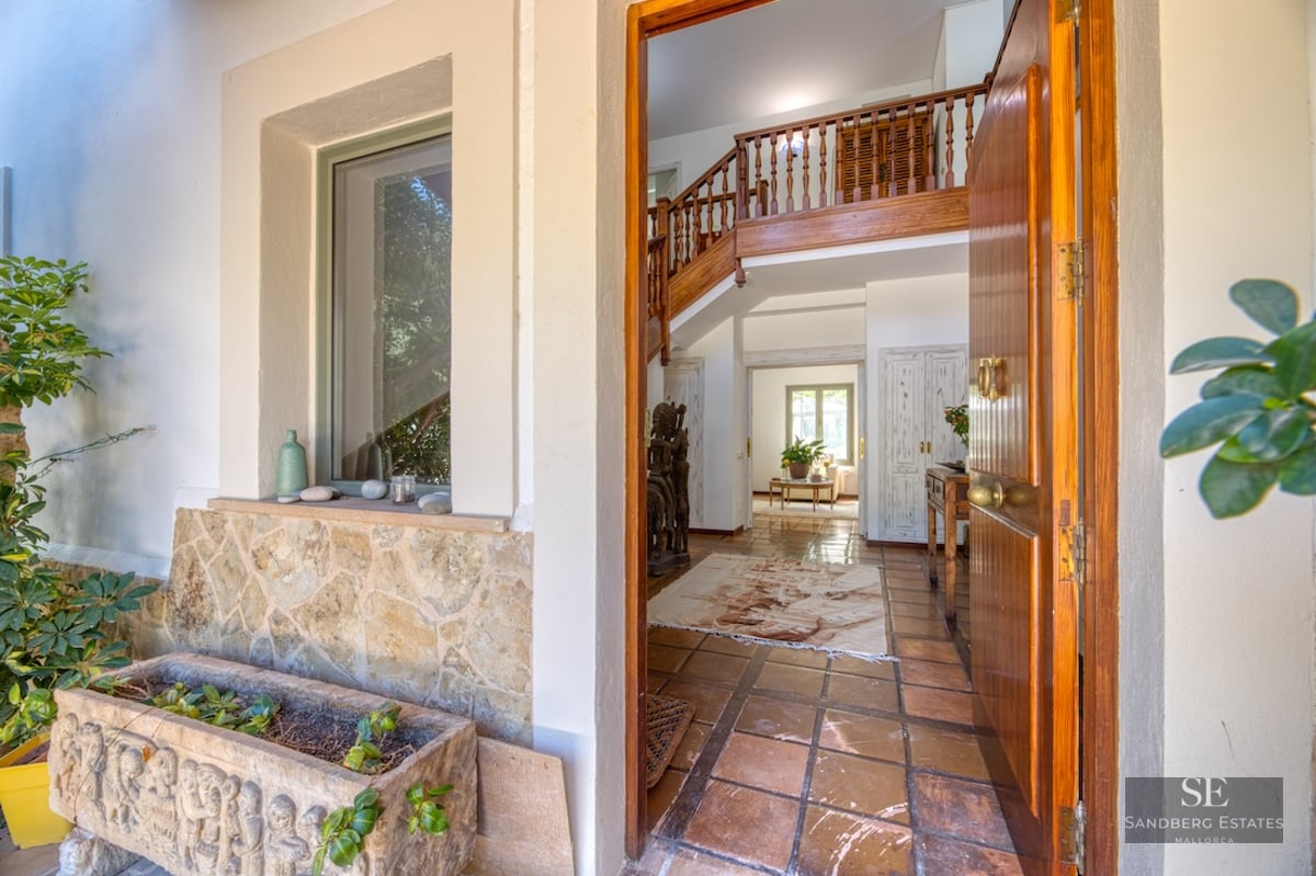 Open wooden door looking into a Mediterranean hallway with terracotta tiles and a wooden staircase.