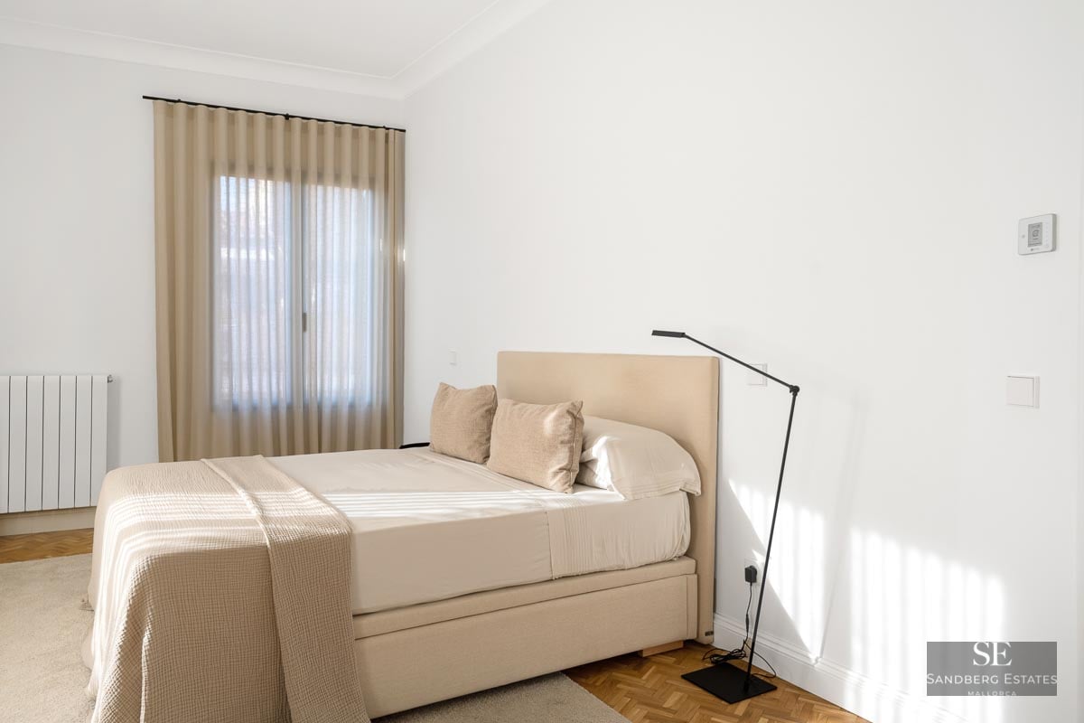 Bright minimalist bedroom with a beige upholstered bed, white walls, and warm wooden parquet flooring.