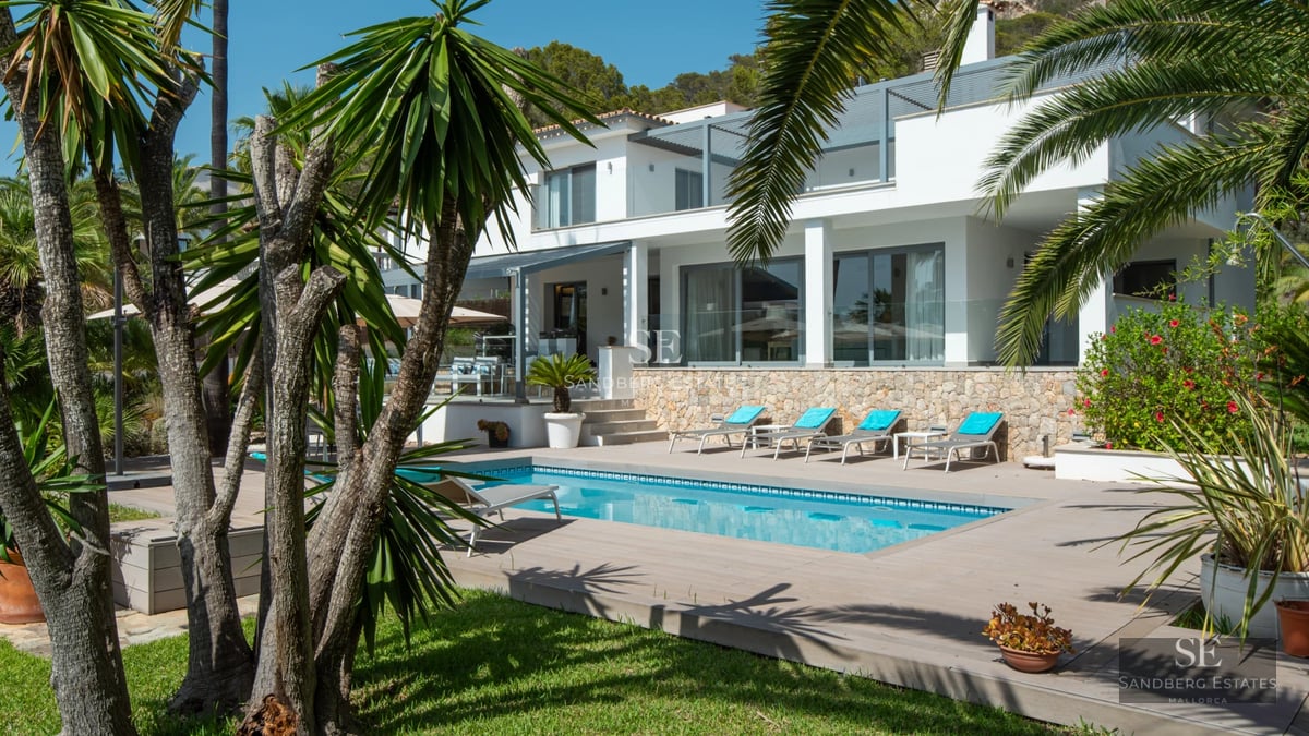 Large swimming pool surrounded by lush greenery, with a terrace and views of a modern and minimalist design house.