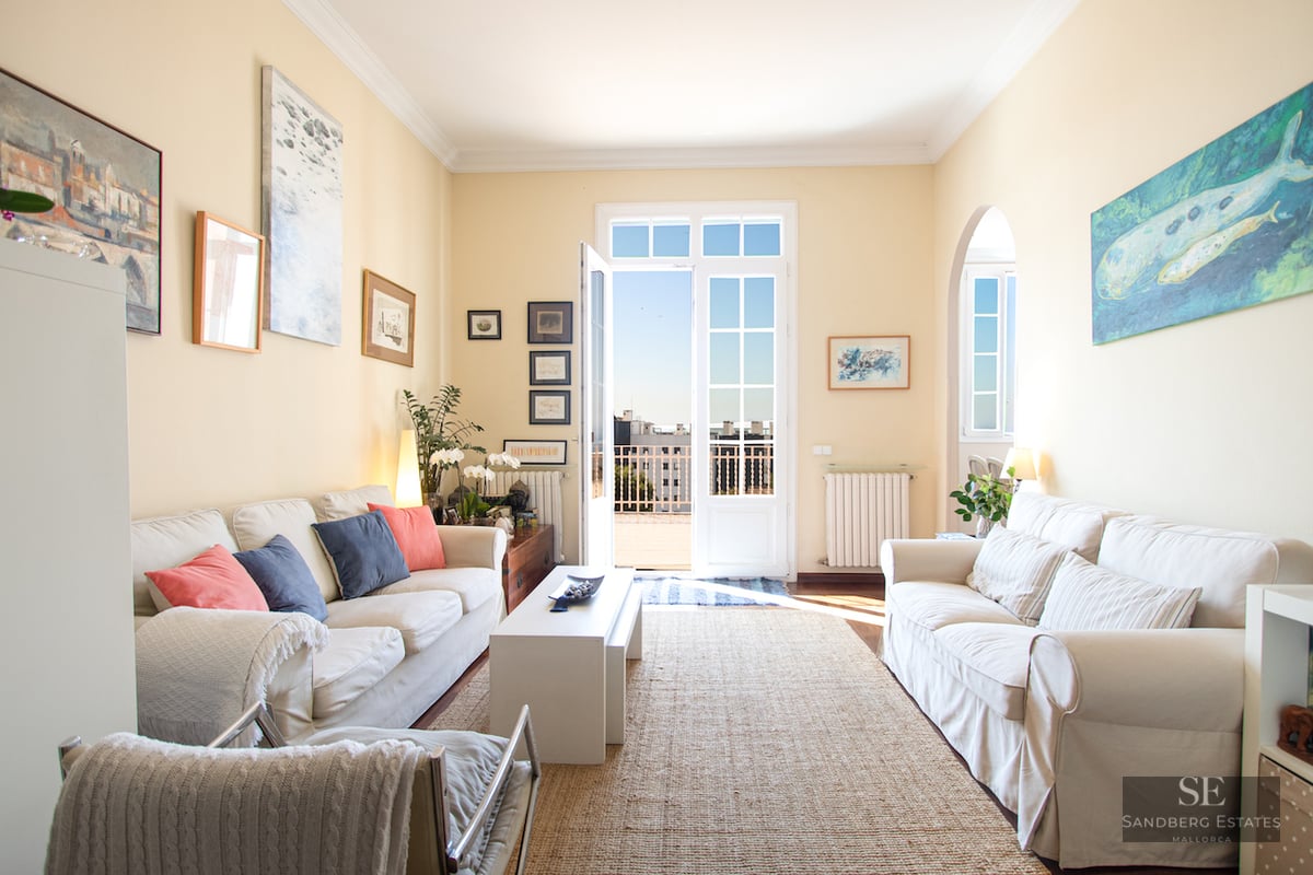 Sun-drenched living room with white sofas, a jute rug, and French doors leading to a balcony with city views.