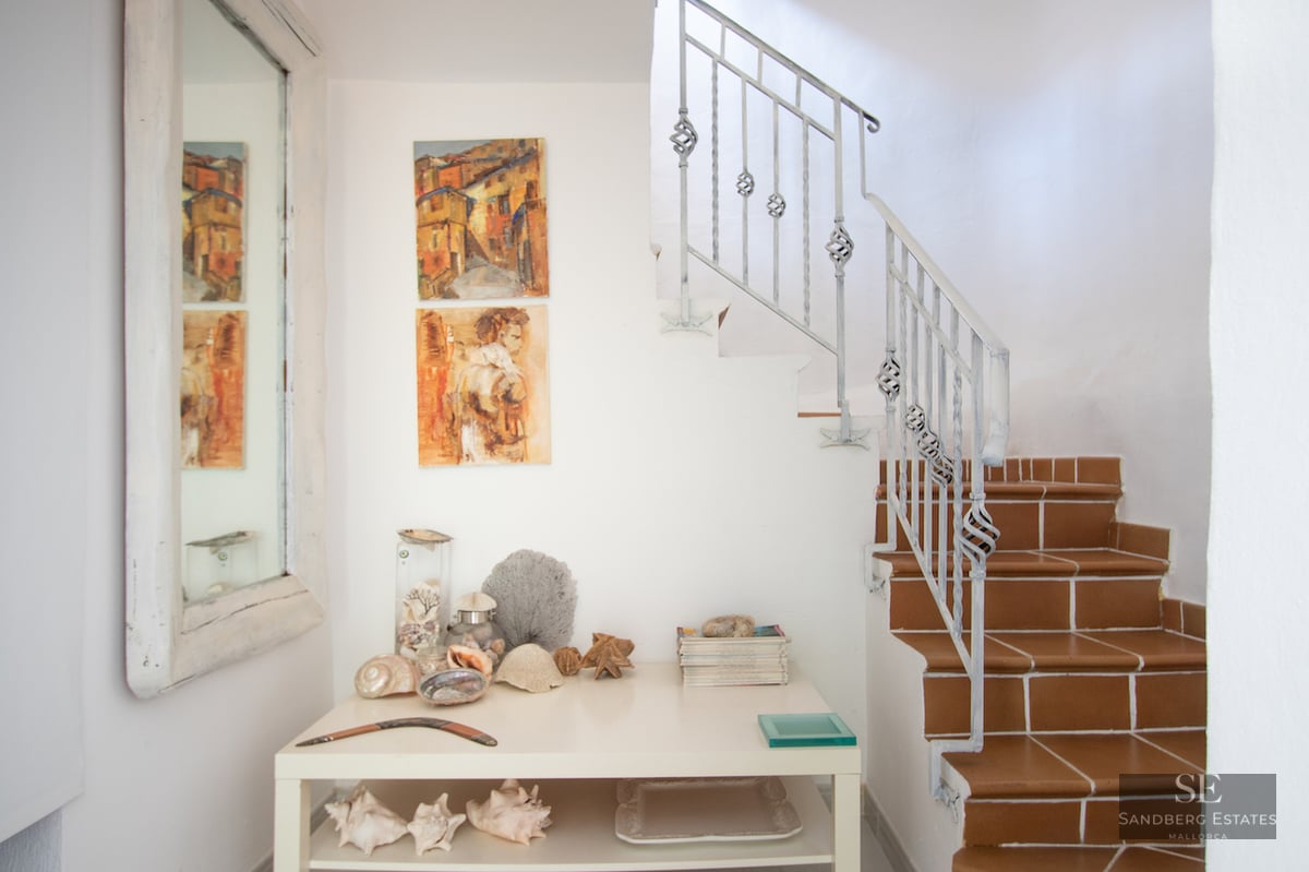 A white hallway featuring terracotta tiled stairs, a wrought iron railing, and a console table with coastal decor.