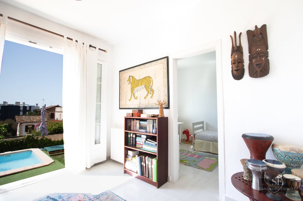 Bright interior with a bookshelf, tribal masks, and an open door leading to an outdoor swimming pool.