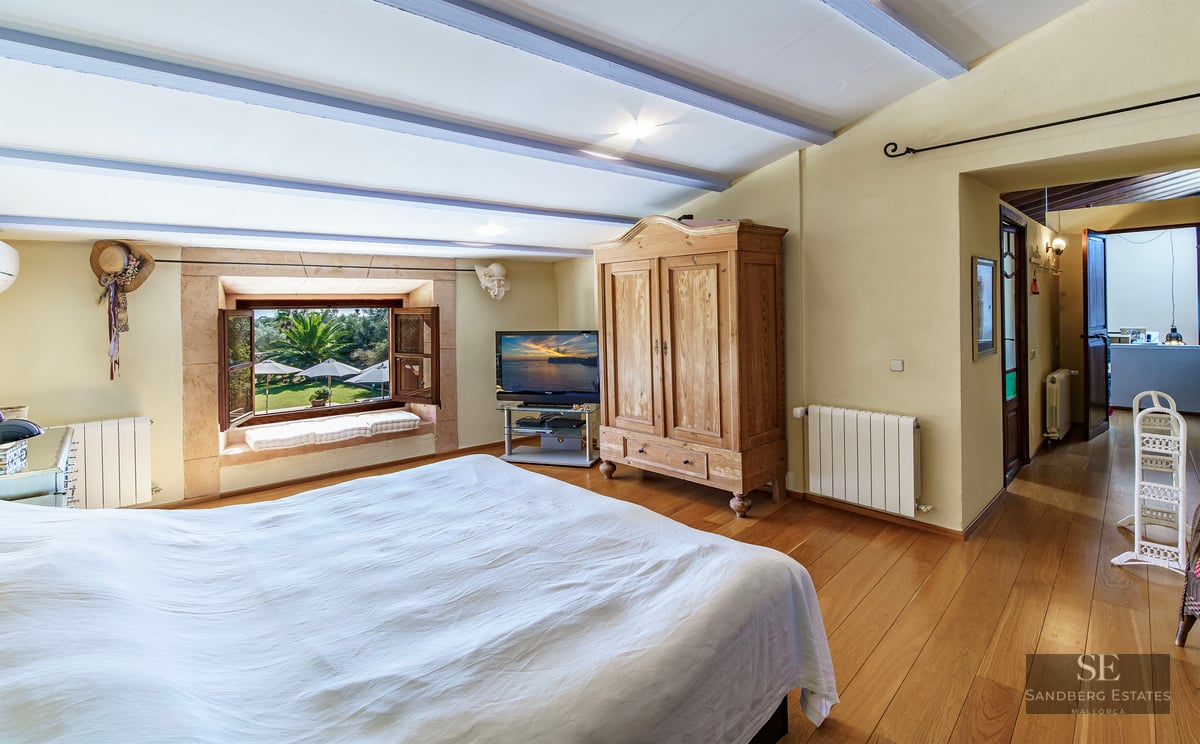 A cozy bedroom with wooden floors, a large bed, rustic armoire, and a window overlooking a lush garden.