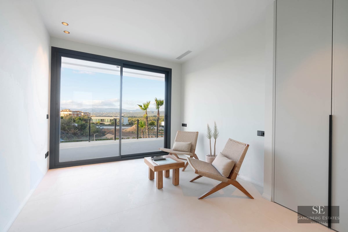 Minimalist white room featuring two design chairs, a wooden table, and large sliding glass doors overlooking a landscape.