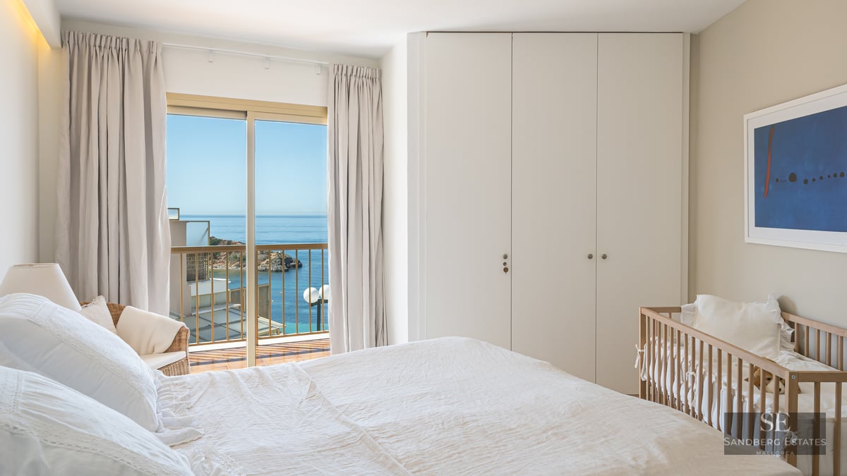 Bright bedroom with white bedding, wooden crib, built-in wardrobe, and sliding glass door opening to a sea view.