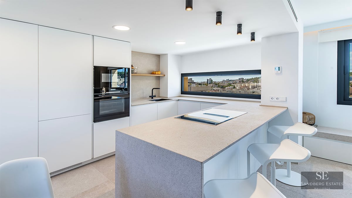 Minimalist white kitchen with large stone island, integrated appliances, and a panoramic window overlooking a city.