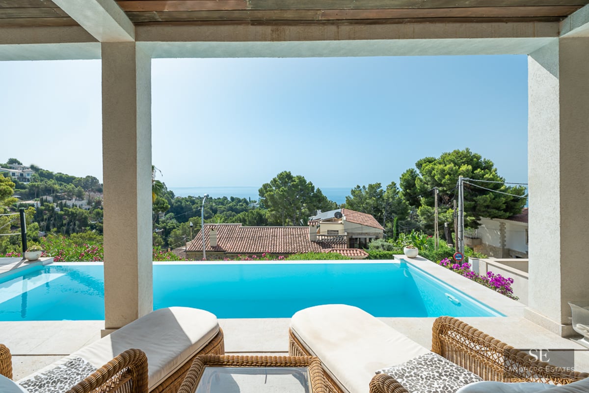 View from a shaded terrace overlooking an infinity pool towards the Mediterranean Sea and lush green hills.