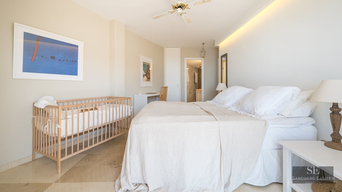 Bright bedroom featuring a large bed, wooden baby crib, workspace, and polished marble flooring.
