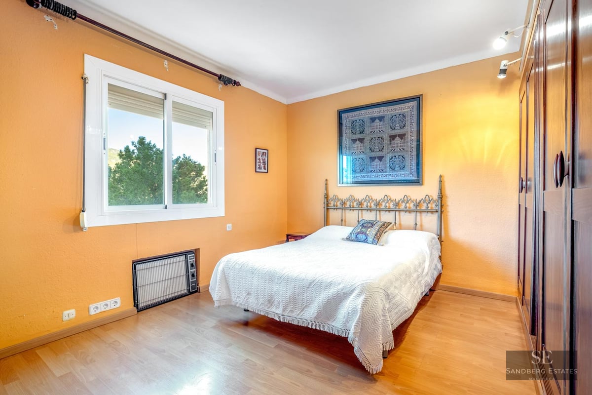 Bedroom with orange walls, white bed, ornate metal headboard, large wooden wardrobe, and window view of trees.