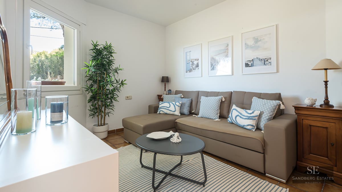A bright living room featuring a taupe L-shaped sofa with blue cushions, a striped rug, and coastal-themed wall art.